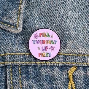 Macro shot of a purple enamel pin with Fill Yourself Up First text on denim jacket