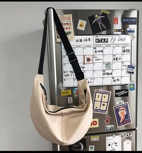 Beige nylon crescent crossbody bag with adjustable black strap shown on a silver fridge