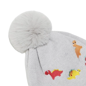 Detail of grey knitted hat featuring fluffy faux fur pompom and colorful dinosaur embroidery