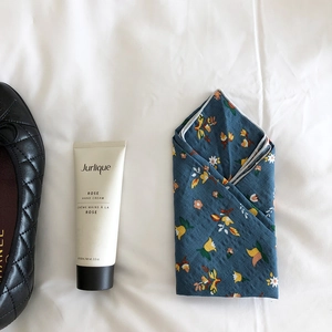 Flat lay of a floral cotton scarf beside a rose hand cream and quilted shoe on white linen