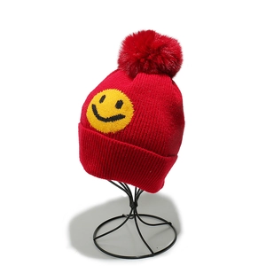 Close-up of red ribbed knit beanie featuring a yellow smiley face patch and faux fur pompom