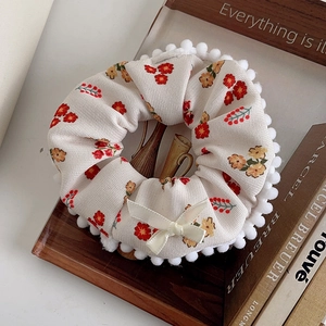 Detail of plush cream scrunchie with floral pattern, white pom-pom trim and bow