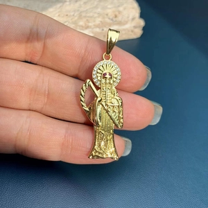 Gold Santa Muerte pendant with white crystals and ruby eyes held by hand on blue leather