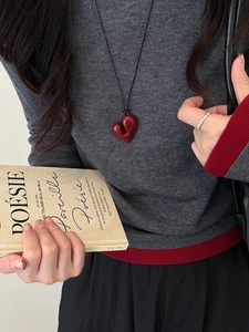 Dettaglio di una collana con ciondolo a cuore rosso 3D e cordino nero su un maglione grigio