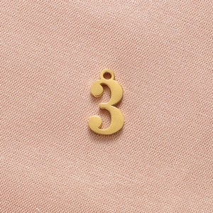 Detail of a matte gold-finished number 3 pendant with a hanging loop on fabric