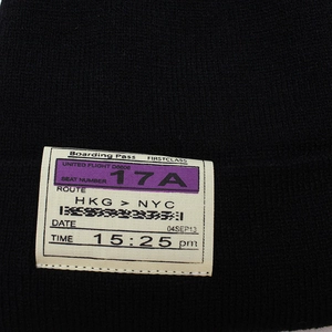 Close-up of black knitted beanie featuring a white boarding pass style woven patch detail