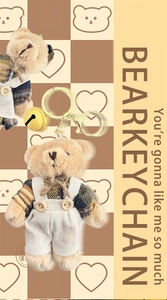 Close-up of plush bear keychain with brown plaid shirt, white overalls and yellow bell