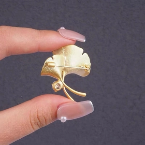 Broche feuille de ginkgo dorée avec perle et cristal tenue en main sur fond gris sombre