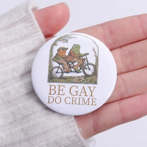 Close-up of a Frog and Toad tin badge pinned on a sweater with Be Gay Do Crime text