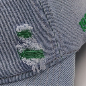 Detail of distressed light blue denim baseball cap featuring green embroidery and frayed rips