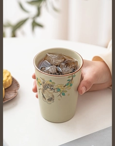 Detail of hand holding a beige ceramic cat mug with ice and tea on a white table