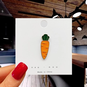 Detail of a cute orange carrot enamel pin with green leaves held by fingers