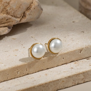 Close-up of elegant white pearl stud earrings on gold base against a beige stone surface