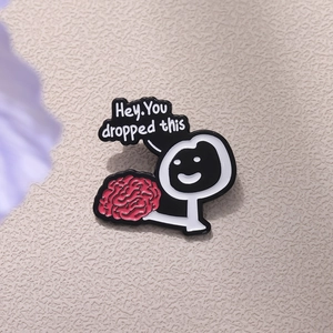 Funny cartoon brain enamel pin with 'Hey, You dropped this' text on beige background
