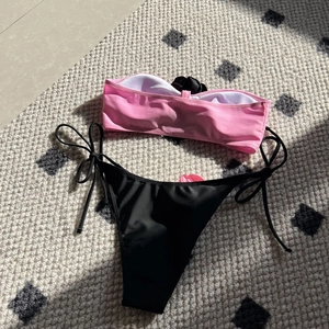 Flat lay detail of pink bandeau bikini top and black side-tie bottoms on a woven rug