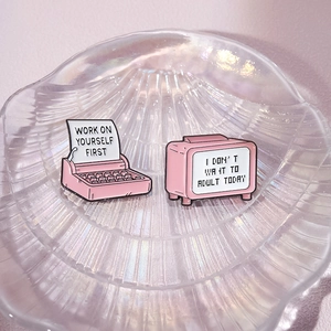Pink enamel pins set with typewriter and retro TV shapes on a decorative shell tray