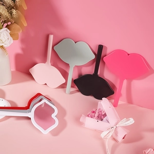 Lip-shaped handheld makeup mirrors in pink, grey, and black on a minimalist vanity