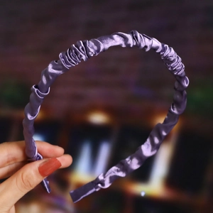Detail showing hand holding a glossy purple pleated satin headband in a soft interior setting