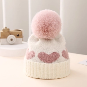 Detailed close-up of ivory knitted baby hat with pink heart patterns and large faux fur pompom
