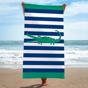 Blue and white striped beach towel with green cartoon crocodile print shown on beach