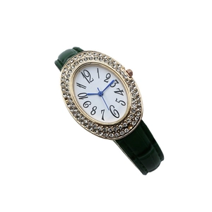 Montre ovale dorée élégante avec lunette en strass et bracelet en cuir vert foncé sur blanc