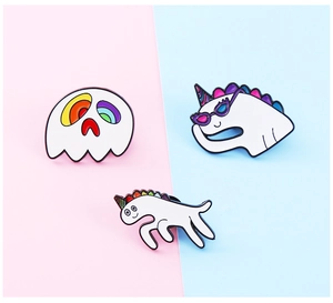Detail of three white enamel pins featuring cartoon ghost, unicorn, and pony on blue-pink ground