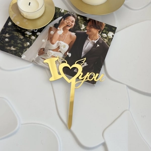 Close-up of golden acrylic I Love You cake topper with winged heart on white surface