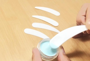 Close-up of white curved spatula scooping blue facial cream from a clear glass jar