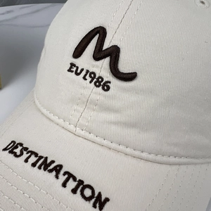 Detail of embroidered letters M and EU 1986 on the cream cotton baseball cap