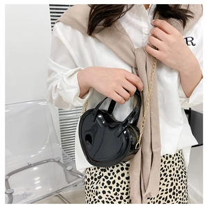 Detail of a heart-shaped black patent leather bag featuring a gold chain strap on a model