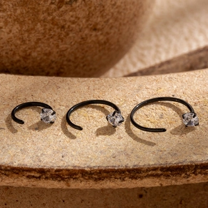 Three black C-shaped G23 titanium nose rings with cubic zirconia on a stone surface