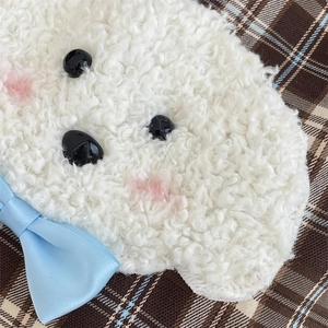 Close-up of white plush dog charm with blue bow on brown plaid background