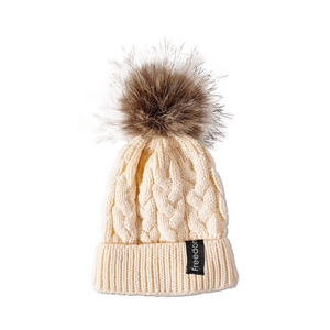 Détail du bonnet tricoté crème pour bébé avec torsades, pompon marron et étiquette Freedom