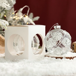 Clear glass Christmas ornament with silver glitter patterns next to white gift packaging