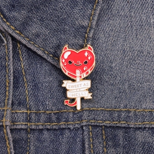 Macro detail of red devil heart lollipop enamel pin with Sweet As Hell text on denim