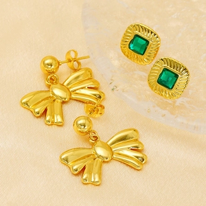 Elegant gold bow earrings and green zircon square studs on silk and glass background