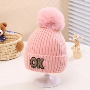 Close-up of pink knitted kids hat with a fluffy pompom and stitched OK patch detail