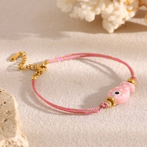 Pink braided fish bracelet with evil eye charm on white sand and coral background
