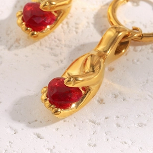 Macro detail of hand-shaped gold earrings holding a red heart gemstone on stone background