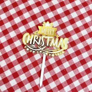 Detailed view of a gold and white Merry Christmas acrylic cake topper on a plaid cloth
