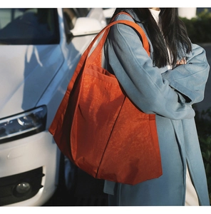 Orange nylon tote bag with pleated texture worn over the shoulder in an urban street setting
