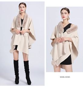 Detail view of beige knitted poncho with thick faux fur collar and pom-pom decorations