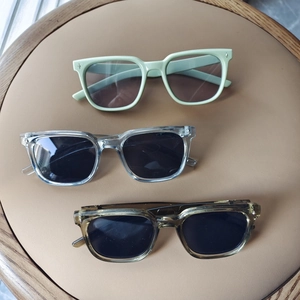 Kids square frame sunglasses in green, blue, and brown displayed on a tan leather base