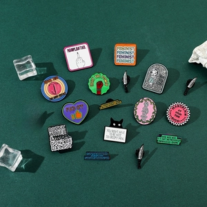 Flat lay of multiple metal enamel pins with feminist and social slogans on green texture