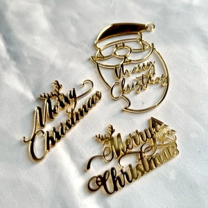 Three gold acrylic Merry Christmas cake toppers with mirror finish on white paper