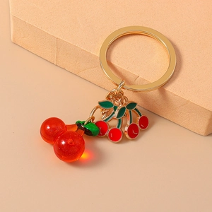 Red resin cherry keychain with golden ring and enamel charms on a warm beige background