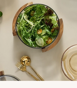 Detail view of a glass salad bowl set with wooden base and golden cutlery in a warm setting