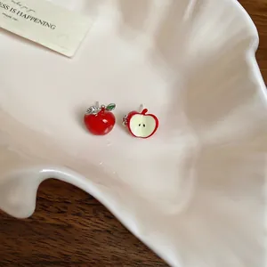 Asymmetric red apple stud earrings with leaf details displayed on a shell ceramic plate