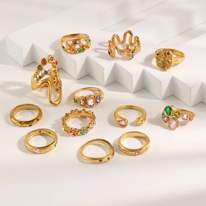 Variety of gold-plated stainless steel rings with colorful crystals on white geometric stand.