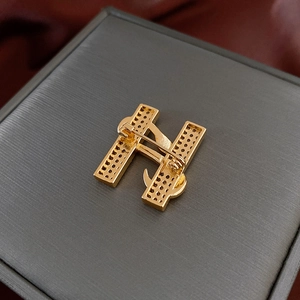 Détail de la broche lettre H dorée montrant l'épingle de sûreté et la finition métal brossé